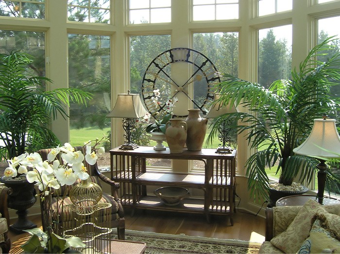 Large sunroom with long side table and lamps along with plant greens everywhere and a large metal clock. 